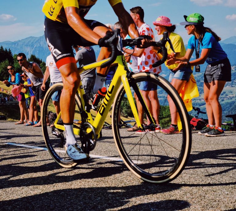 A rider on a yellow BMC bike cycles up Col de Romme in the 2018 Tour de France with a row of supporters cheering in the background.