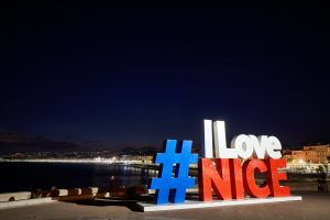 The red, white, and blue "hashtag I Love Nice' sign lit up at night with the city in darkness behind.