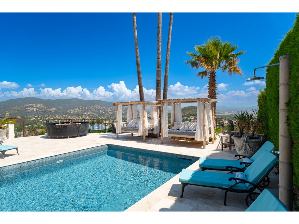 A view of the pool across the landscaped terrace of sun loungers and day beds, palm trees and clear blue sky with views across the countryside of the French Riviera in the background.